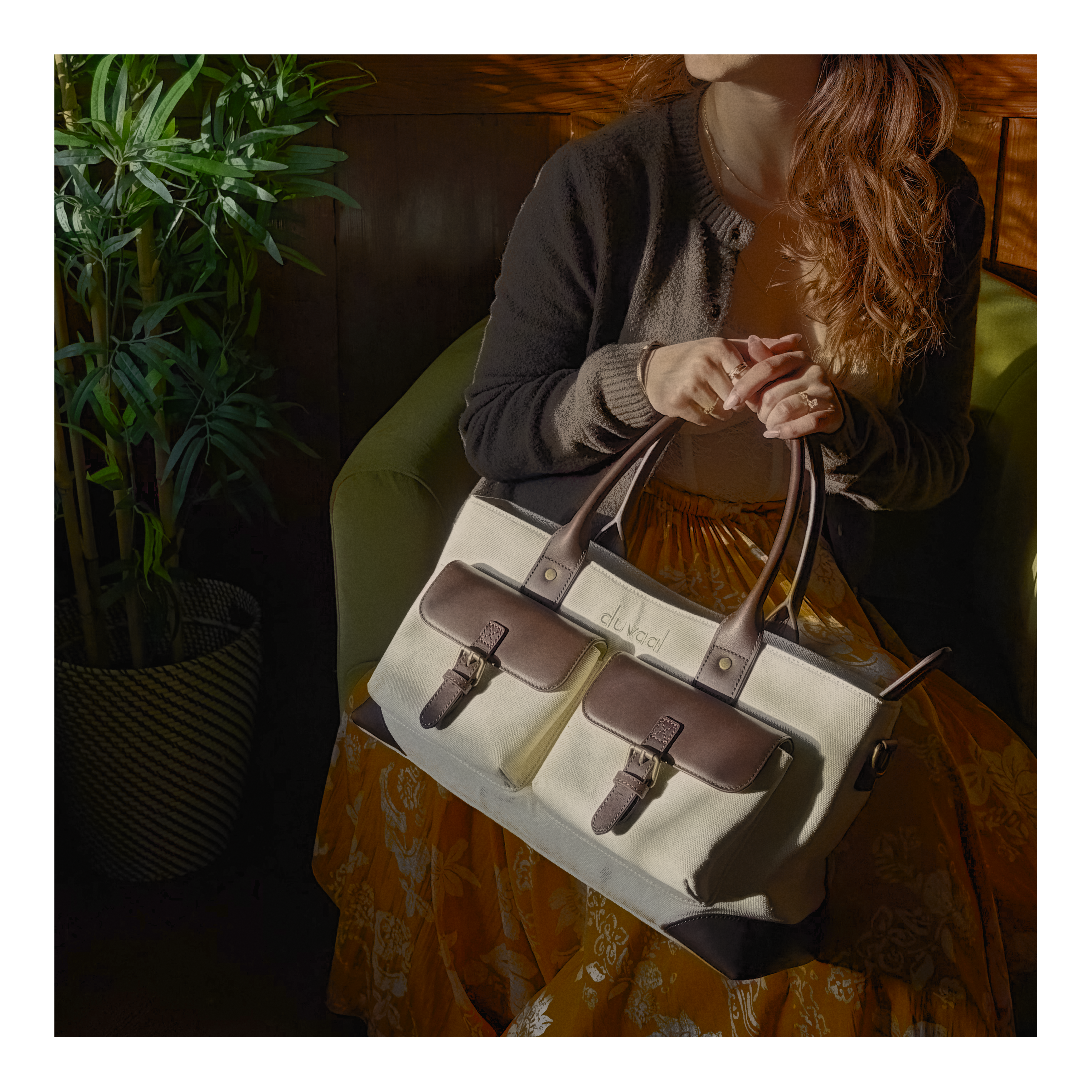 Woman sitting indoors holding a beige canvas tote bag with brown leather accents and pockets